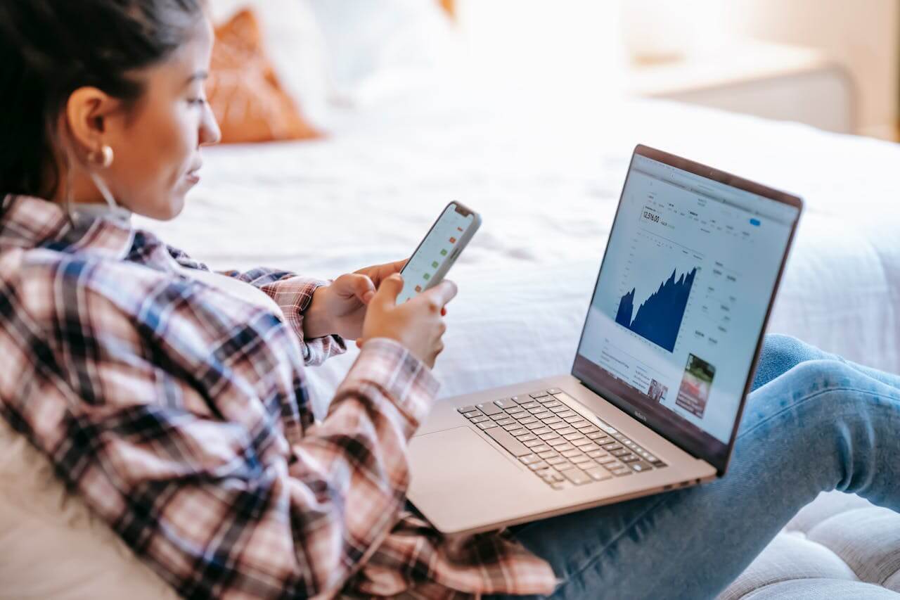 A young girl in the check-style shirt with the blue jeans using her phone while also working on a laptop to provide financial analyst services