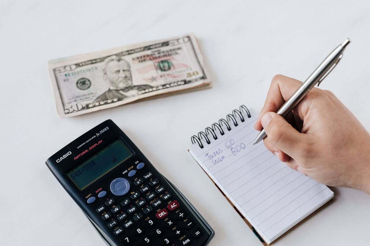 A person writing on a notebook and a calculator and money placed beside the notebook